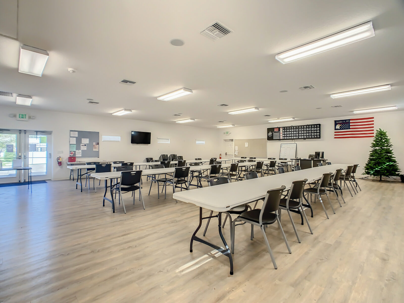 A spacious, well-lit classroom or meeting room with several rectangular tables and black chairs. An American flag and a Christmas tree are in one corner, and there are bulletin boards and a TV on the walls.
