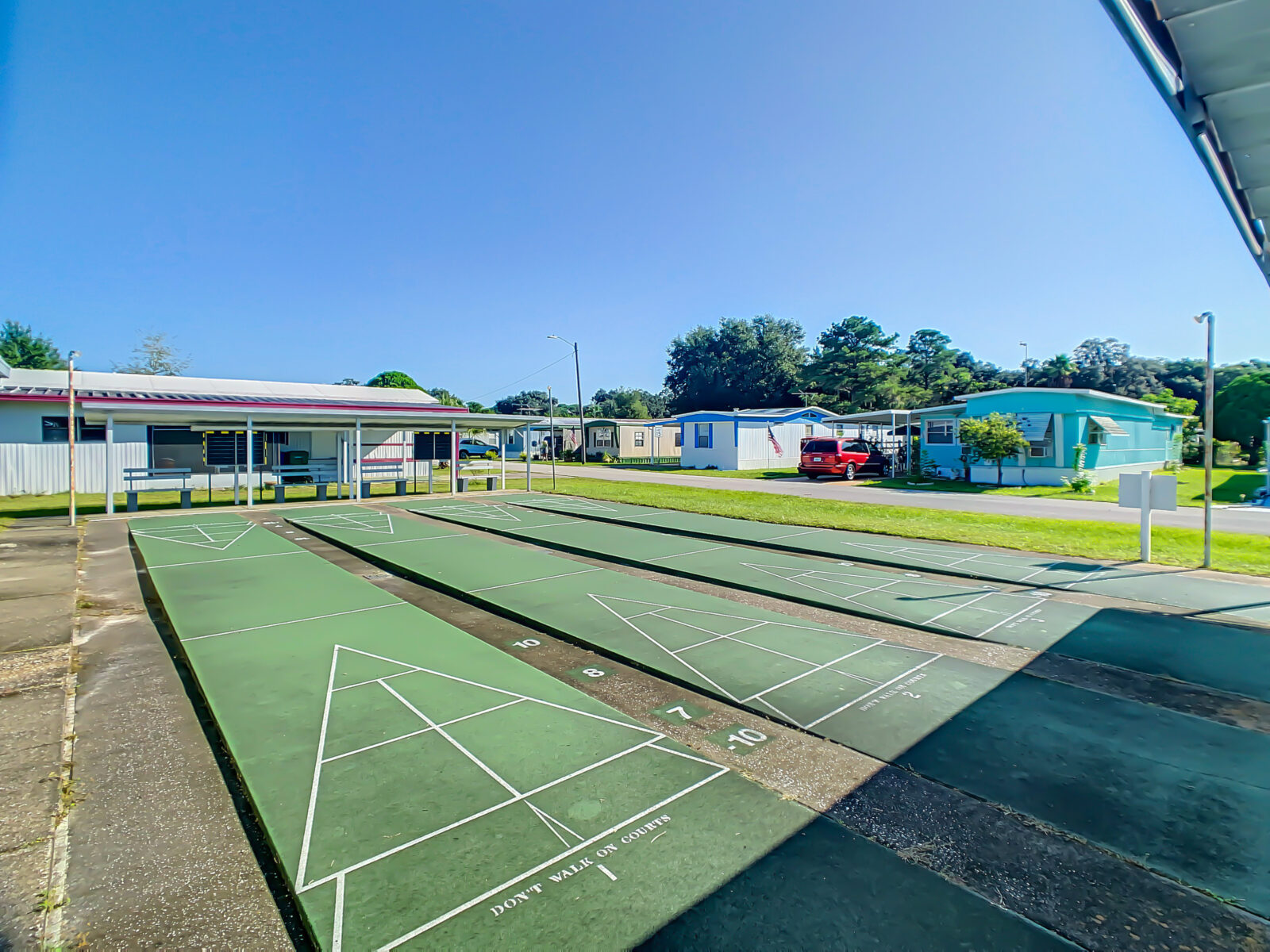 A sunny outdoor shuffleboard court with three green lanes is shown, surrounded by a few mobile homes and a red car parked in the background. Blue sky and trees complete the setting.