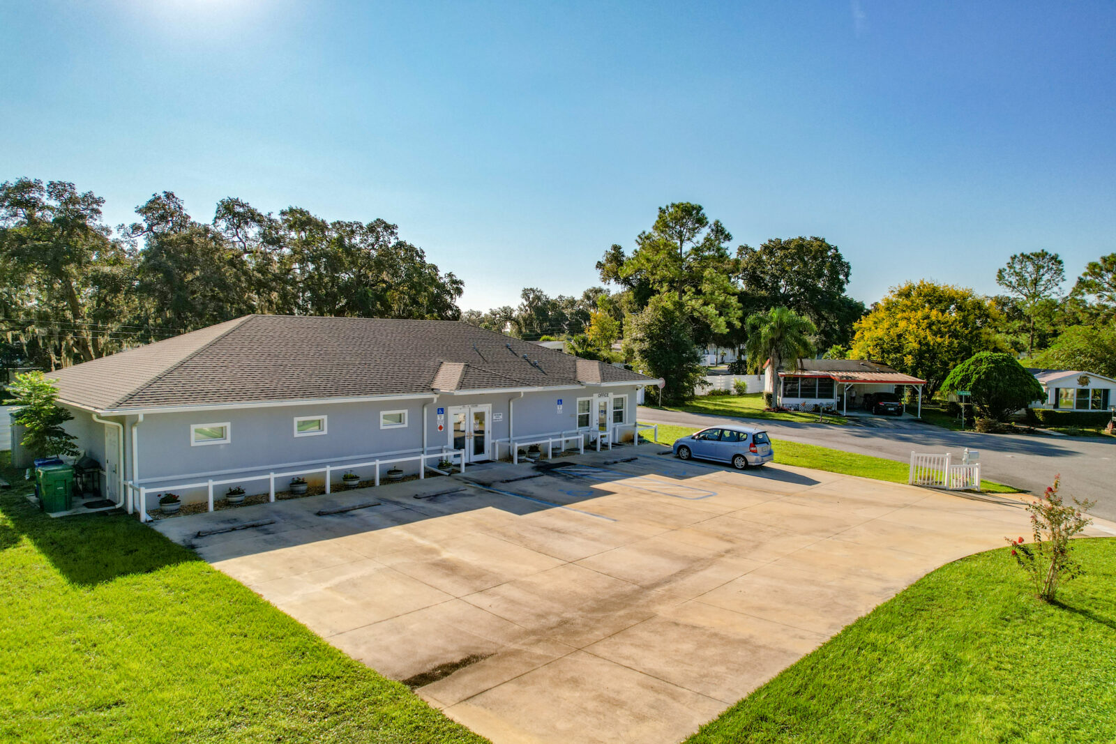 A single-story gray building with a wide driveway and parking area sits on a sunny day. One car is parked outside, and trees and houses are visible in the background. The sky is clear and blue.