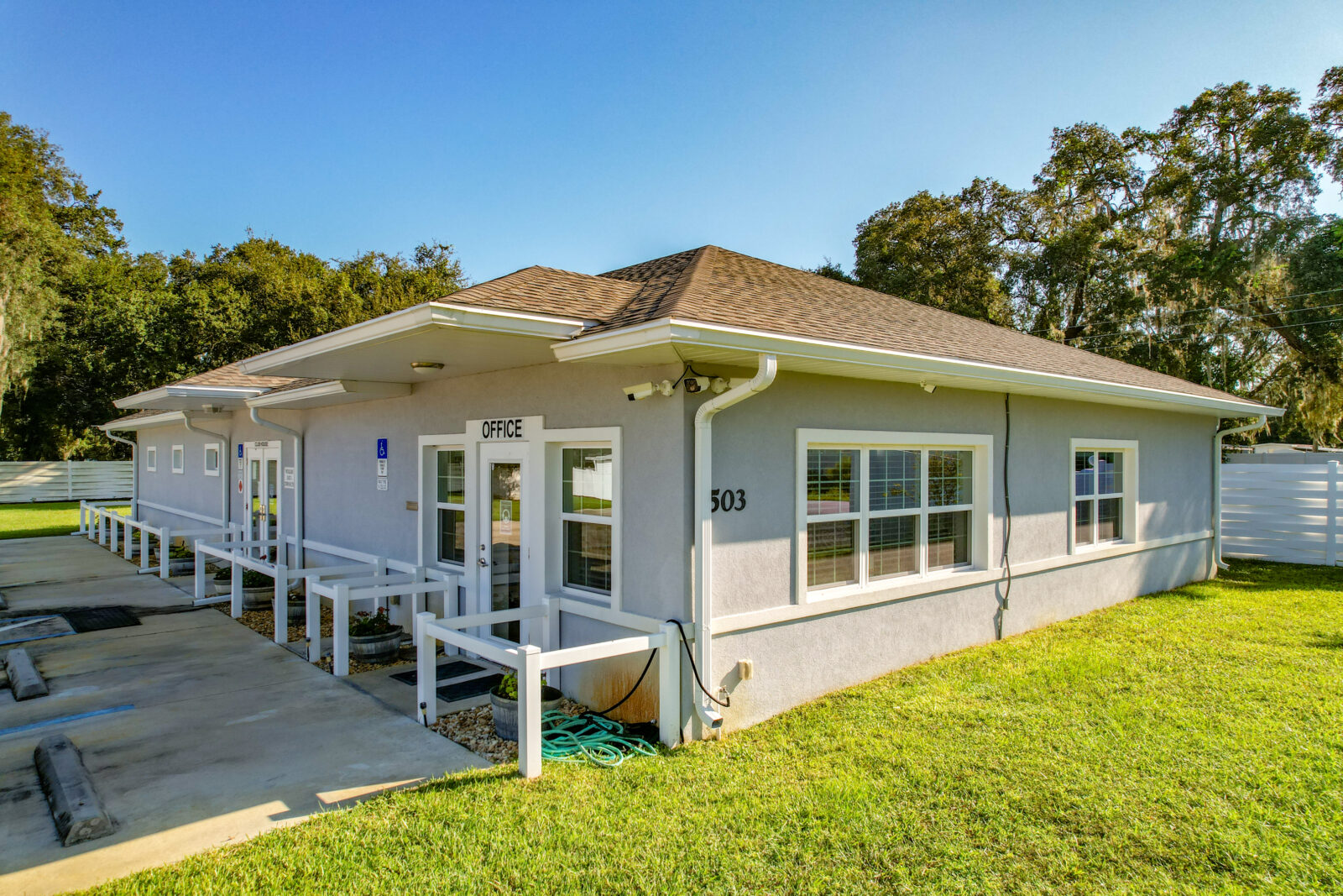 A single-story light gray building marked Office with large windows, white railings, a sloped roof, and a grassy yard, set against a backdrop of trees and clear blue sky.
