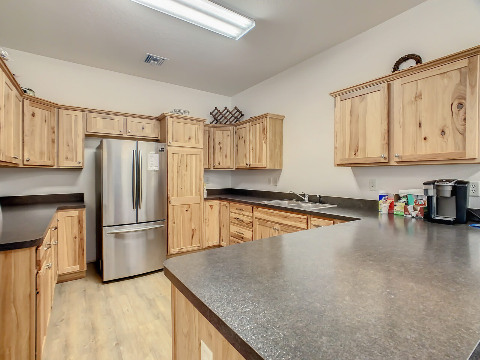 A spacious kitchen with light wood cabinets, black countertops, a stainless steel refrigerator, a sink, coffee maker, and various items on the counter, under bright ceiling lights.