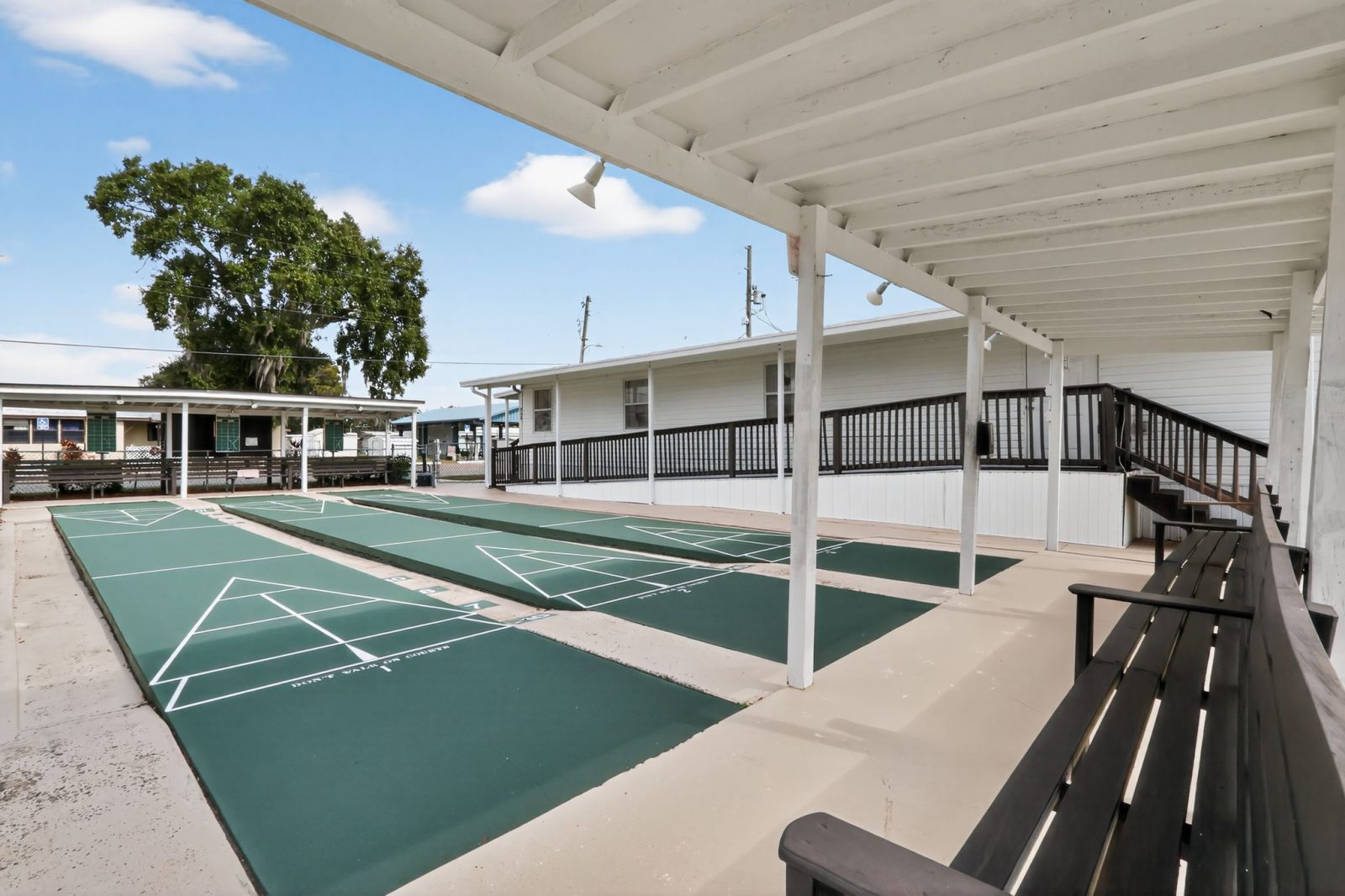 Covered outdoor shuffleboard courts with green playing surfaces, white lines, and benches along the side. A ramp leads to a white building with black railings. Trees and buildings are visible in the background.