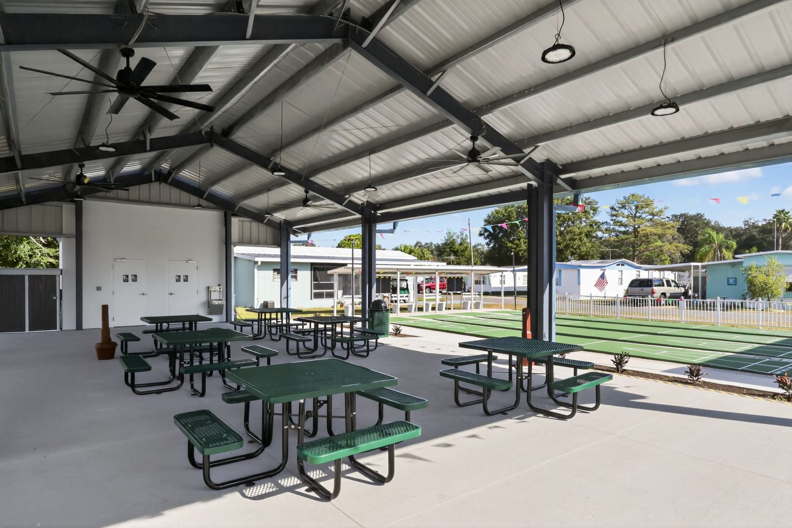 Covered outdoor pavilion with green picnic tables and benches on a concrete floor, ceiling fans above, and shuffleboard courts next to the seating area. Buildings and trees are visible in the background.