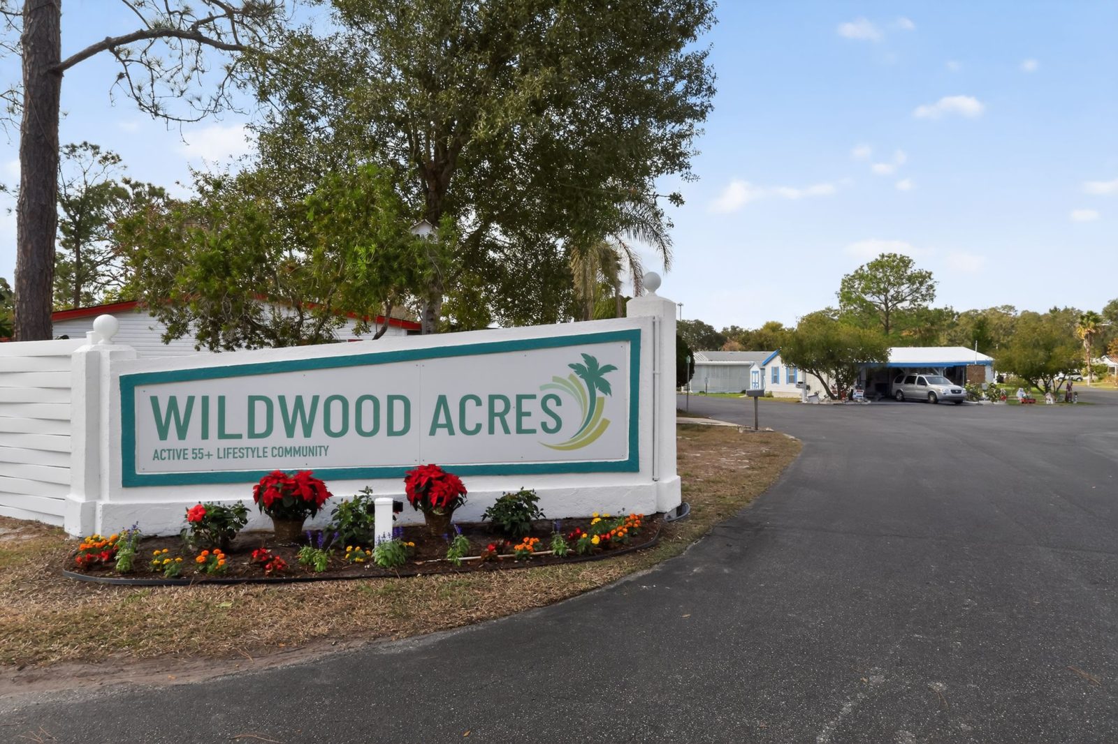 Large white sign reading Wildwood Acres Active 55+ Lifestyle Community stands by the entrance, decorated with red poinsettias. Trees, small white buildings, and a blue sky are visible in the background.