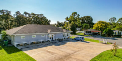 A single-story gray building with a wide driveway and parking area sits on a sunny day. One car is parked outside, and trees and houses are visible in the background. The sky is clear and blue.