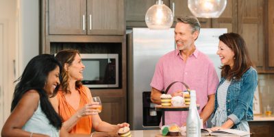 Four adults, two men and two women, smile and chat around a kitchen island with cupcakes and drinks. Pendant lights hang above, and kitchen cabinets and a refrigerator are in the background.